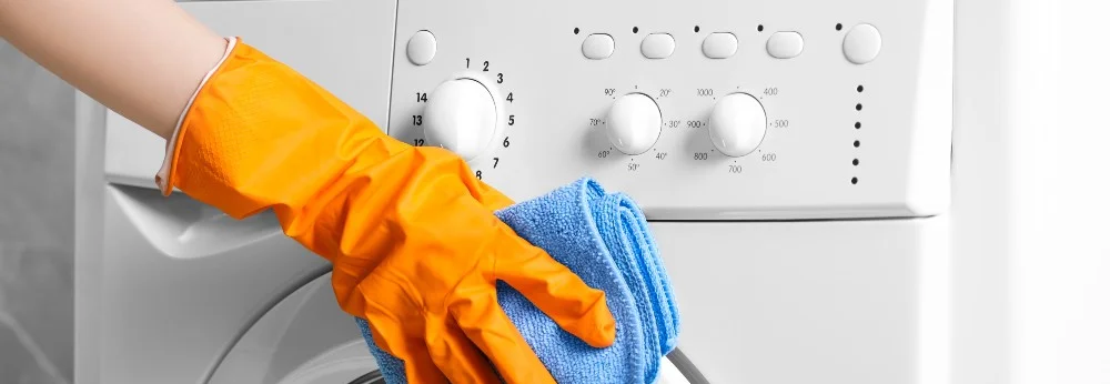 Eine Person mit orangefarbenen Handschuhen reinigt eine Waschmaschine mit einem blauen Lappen.